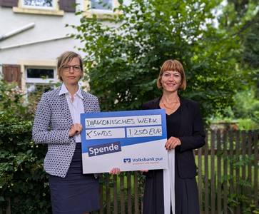 Quelle: Diakonisches Werk Karlsruhe Zwei Damen halten im Rahmen einer Spendenübergabe einen bergroßen Spendenscheck. Sie stehen in einer grünen Umgebung.