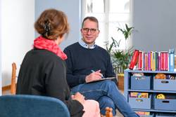 Ein Mann berät eine Frau in einem Raum der Evangelischen Beratungsstelle für Erziehungs-, Ehe- und Lebensfragen in Mülheim an der Ruhr. Der Mann sitzt mit dem Gesicht zur Kamera der Frau gegenüber, die mit dem Rücken zur Kamera sitzt. Er hat kurzes braun-graues Haar und eine Brille. Er trägt ein hellblaues Hemd unter einem dunkelblauen Pullover. Er hat ein Klemmbrett in der Hand und redet gerade. Die Frau hat rotes Haar und trägt einen schwarzen Blazer und eine rotes Tuch.
In dem Raum sind rechts Bücher in einem Regal zu erkennen, sowie diverse Utensilien für Beratungen - zum Beispiel Holzfiguren, ein Plüschtier und Spielzeug.
Im Hintergrund erkennt man eine Pflanze und eine Sitzgruppe mit Kinderstühlen.