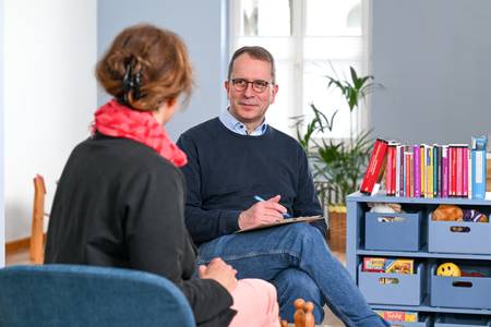 Ein Mann berät eine Frau in einem Raum der Evangelischen Beratungsstelle für Erziehungs-, Ehe- und Lebensfragen in Mülheim an der Ruhr. Der Mann sitzt mit dem Gesicht zur Kamera der Frau gegenüber, die mit dem Rücken zur Kamera sitzt. Er hat kurzes braun-graues Haar und eine Brille. Er trägt ein hellblaues Hemd unter einem dunkelblauen Pullover. Er hat ein Klemmbrett in der Hand und redet gerade. Die Frau hat rotes Haar und trägt einen schwarzen Blazer und eine rotes Tuch.
In dem Raum sind rechts Bücher in einem Regal zu erkennen, sowie diverse Utensilien für Beratungen - zum Beispiel Holzfiguren, ein Plüschtier und Spielzeug.
Im Hintergrund erkennt man eine Pflanze und eine Sitzgruppe mit Kinderstühlen.